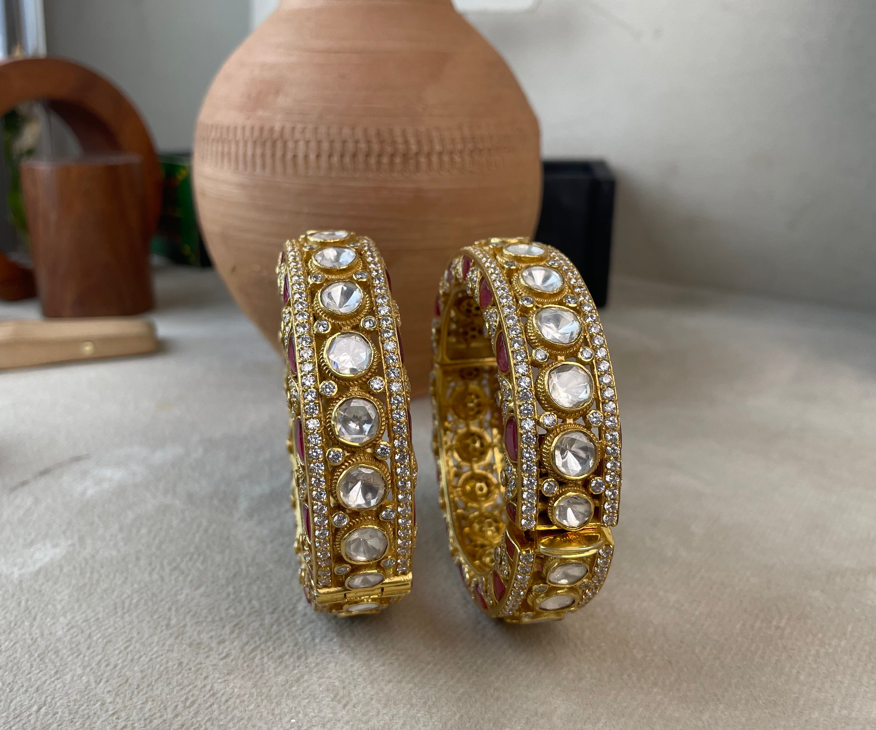 A pair of gold-toned traditional Indian bangles adorned with clear stones, displayed on a neutral surface with a terracotta vase in the background.