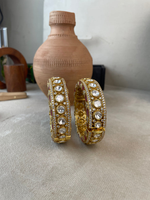 A pair of gold-toned traditional Indian bangles adorned with clear stones, displayed on a neutral surface with a terracotta vase in the background.