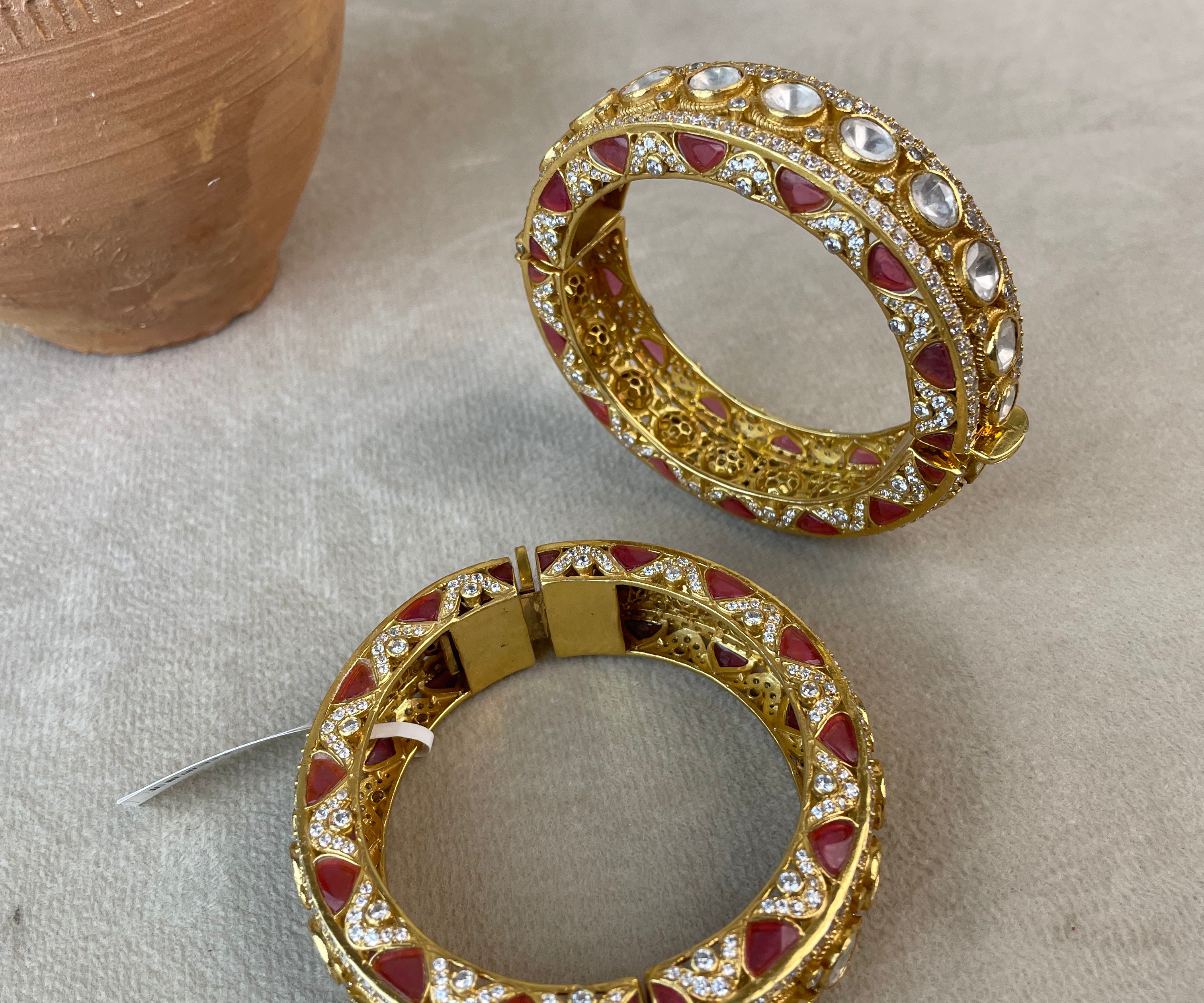 A pair of gold-plated hoop Bangles decorated with red stones and clear crystals, displayed on a neutral beige fabric surface with decorative props.
