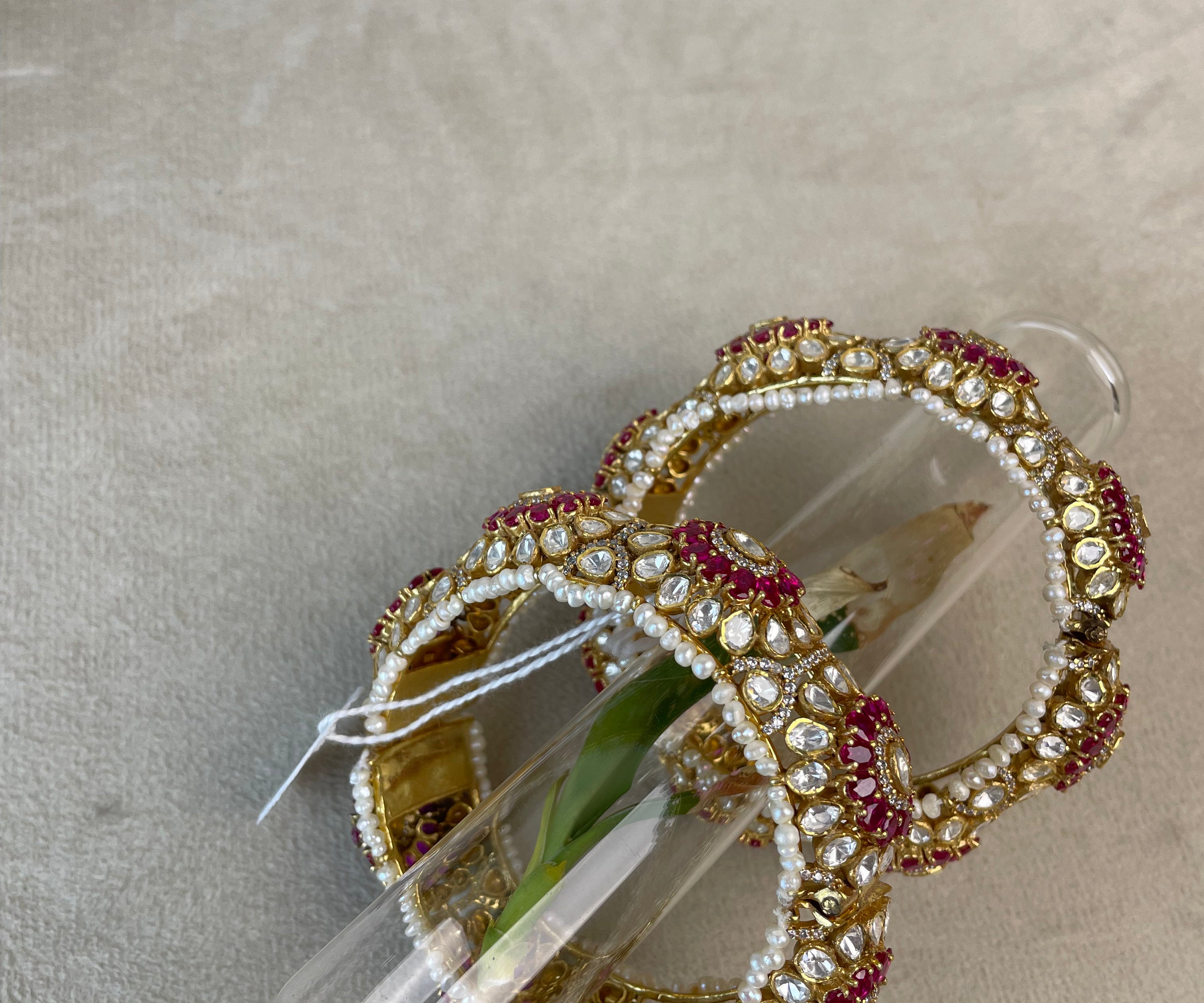 Two ornate bangles decorated with white pearls and red stones, displayed inside clear plastic tubes on a beige textured surface.