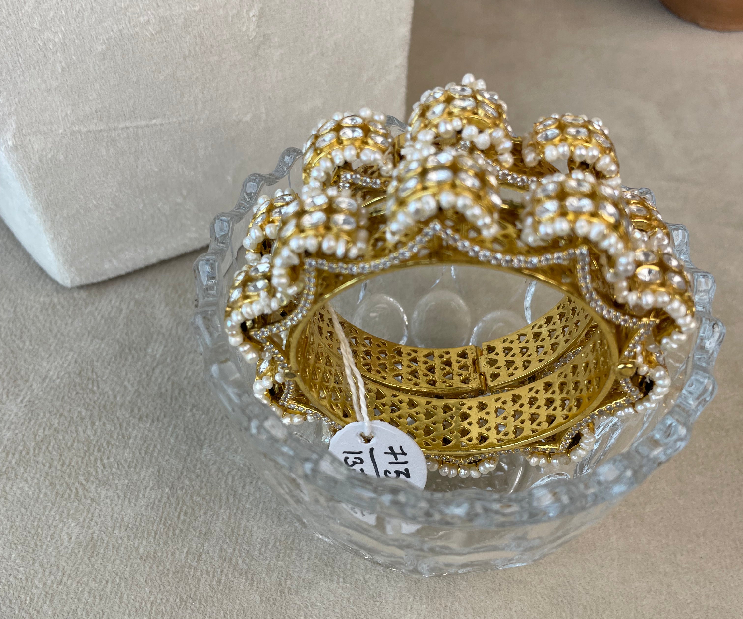 A gold-toned bangle bracelet with intricate pearl beadwork and pavé detailing, displayed inside a clear glass dish on a beige fabric surface.