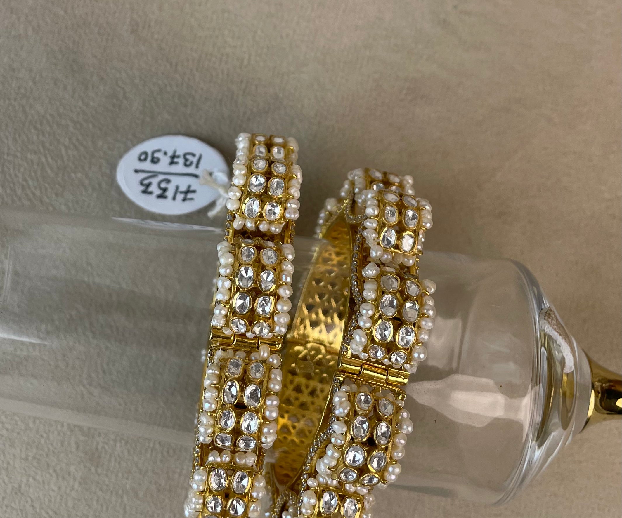 A pair of wide gold bangles heavily embellished with rows of white faux pearls and clear rhinestones, displayed on a clear glass stand over a beige surface.