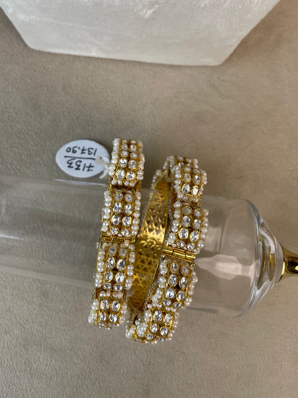 A pair of wide gold bangles heavily embellished with rows of white faux pearls and clear rhinestones, displayed on a clear glass stand over a beige surface.
