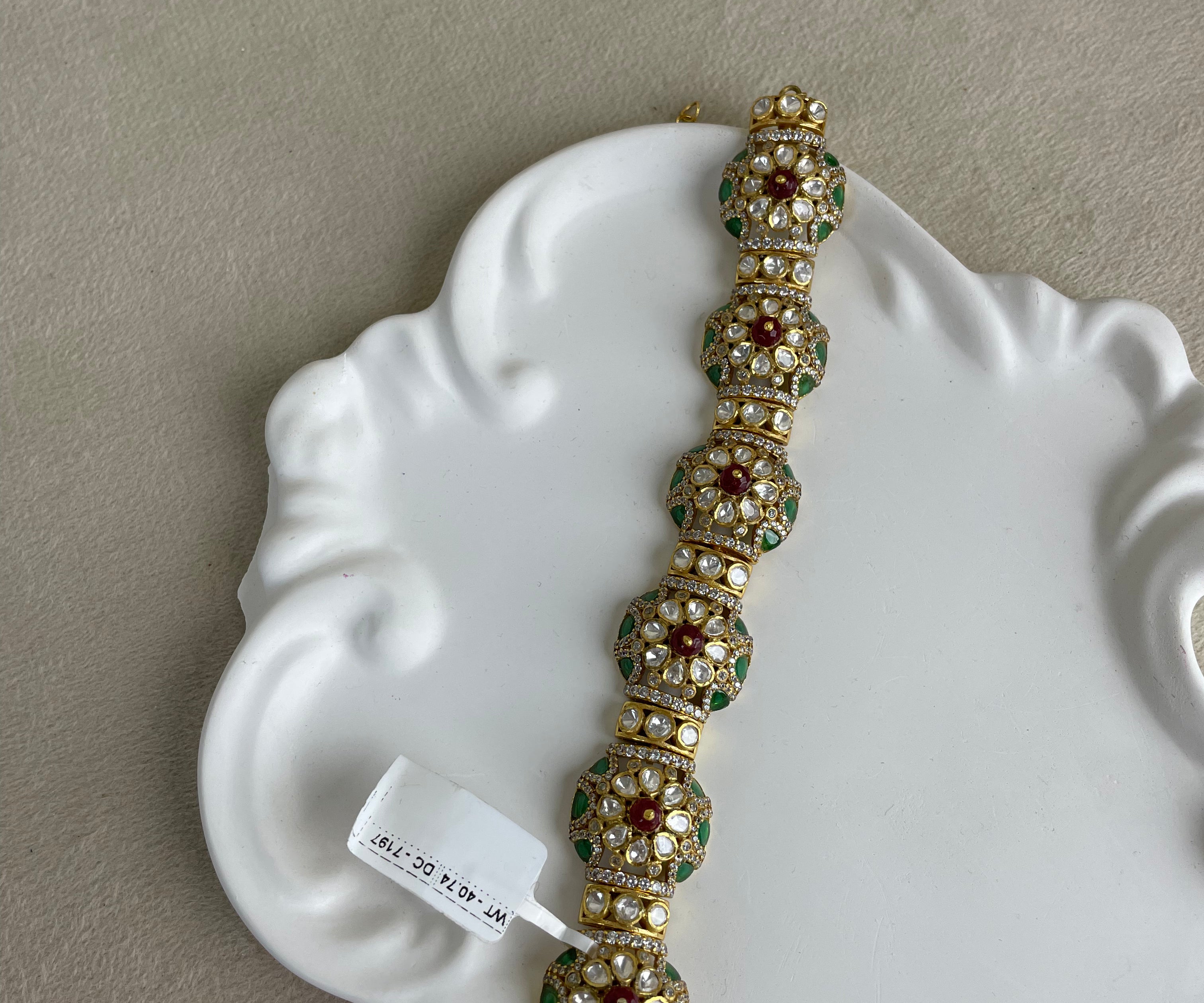 A gold-plated traditional Indian-style bracelet with green and red enamel floral motifs and white stones, displayed on a white decorative ceramic dish.