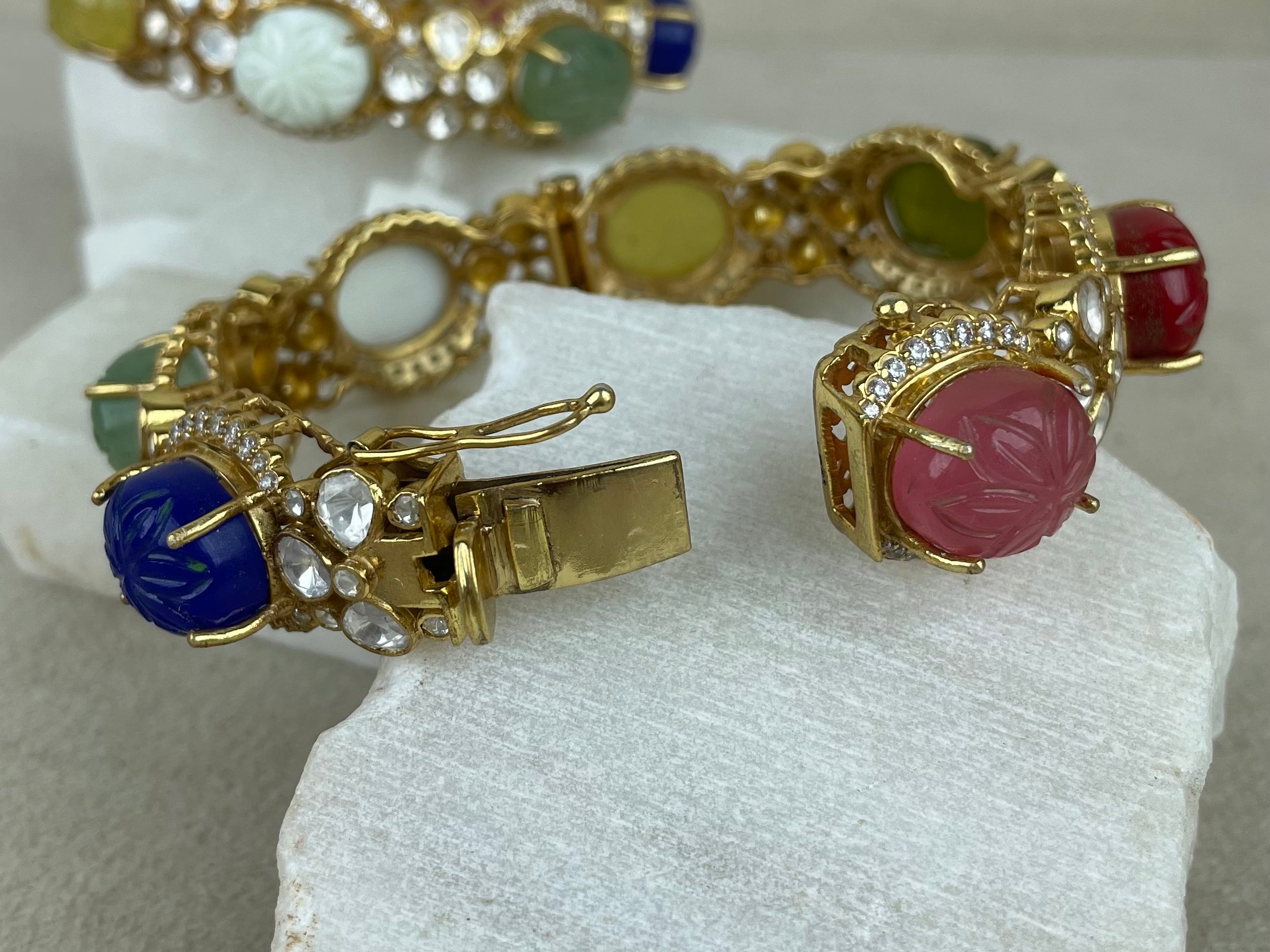 A gold-tone bangle with colorful carved cabochon stones and clear rhinestone accents, displayed on a white stone slab with a second matching bangle in the background.
