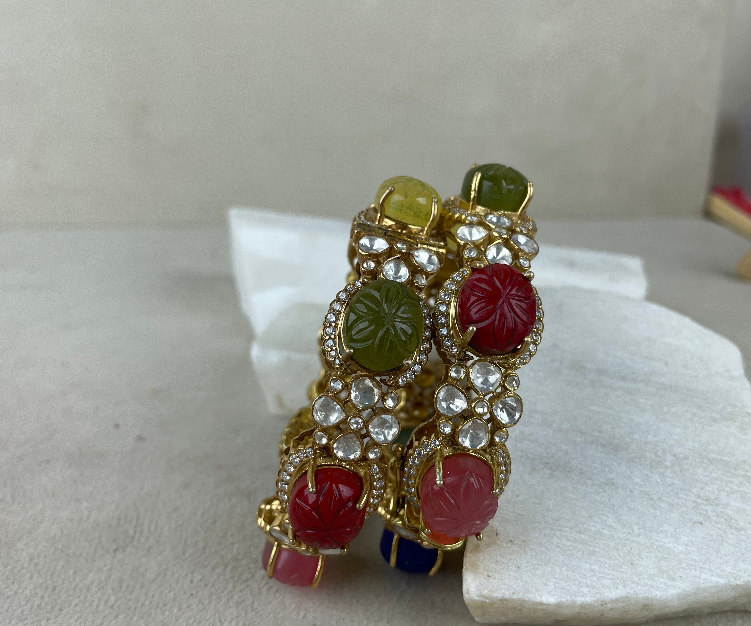 A pair of gold-plated bangles decorated with red, green, and yellow carved stone cabochons and clear rhinestones, displayed on a white stone prop against a neutral beige background.