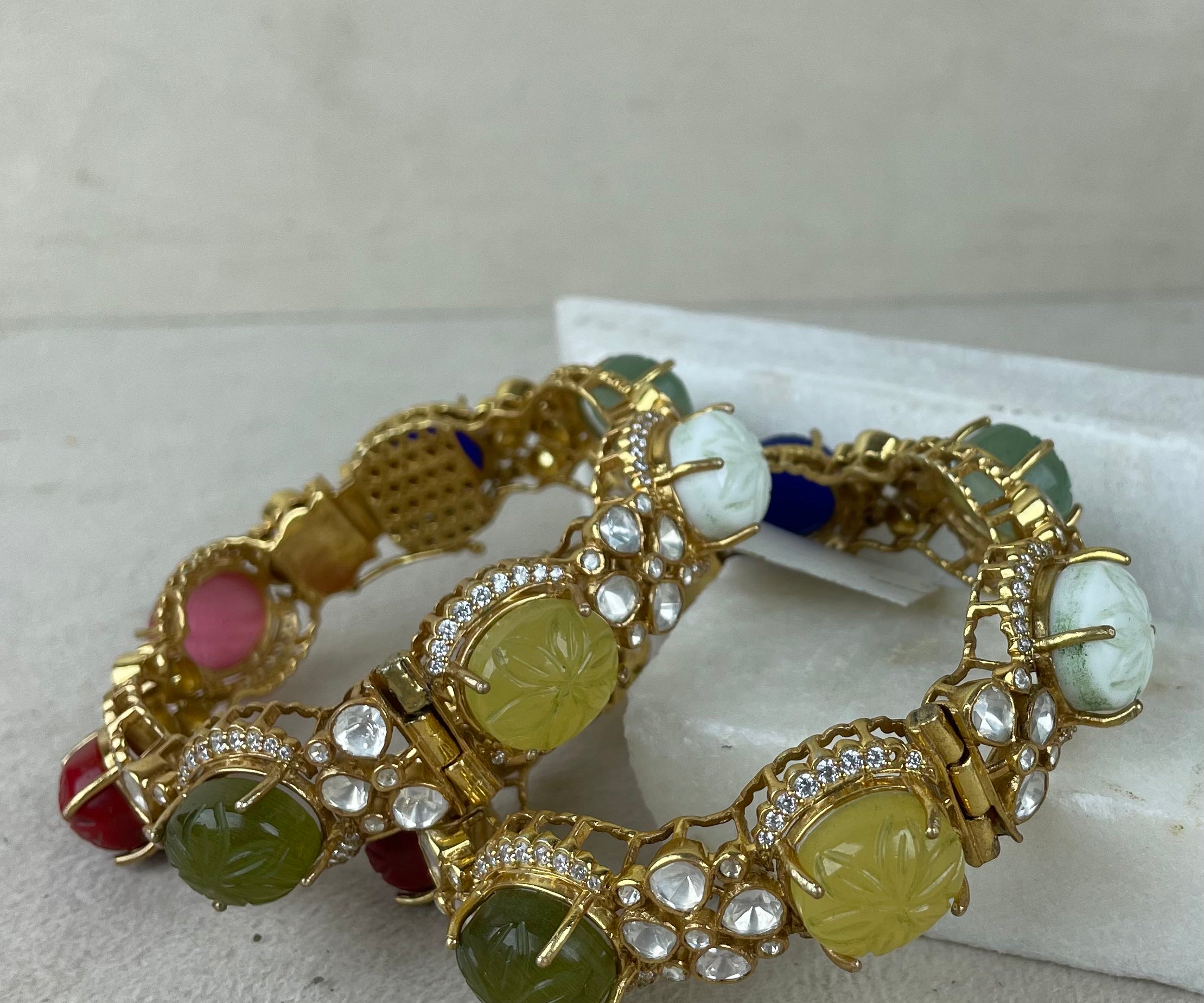 A pair of gold-plated bangles decorated with carved green, yellow, pink, and white stones, set with clear rhinestones, resting on a white display block against a neutral beige background.