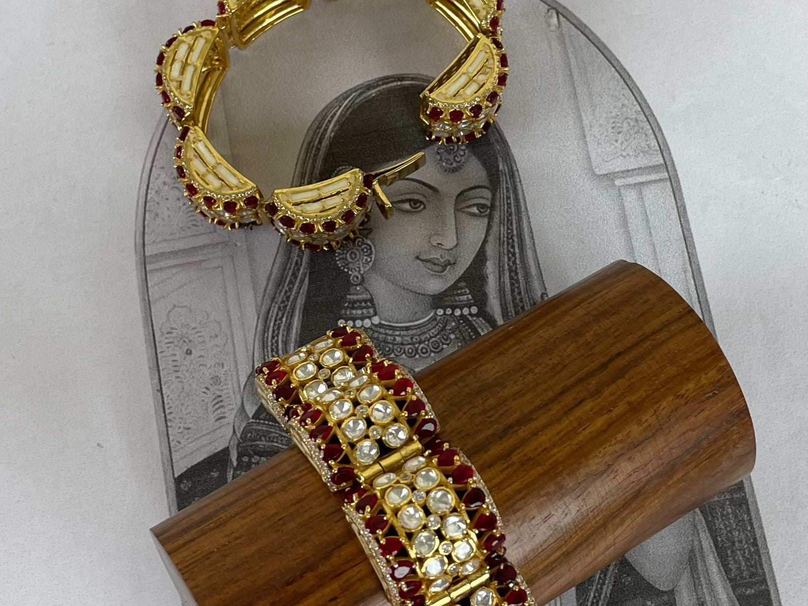 Two ornate gold-toned bangles with red and white stone embellishments, displayed on a wooden stand and a printed card over a light gray surface.