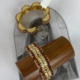 Two ornate gold-toned bangles with red and white stone embellishments, displayed on a wooden stand and a printed card over a light gray surface.