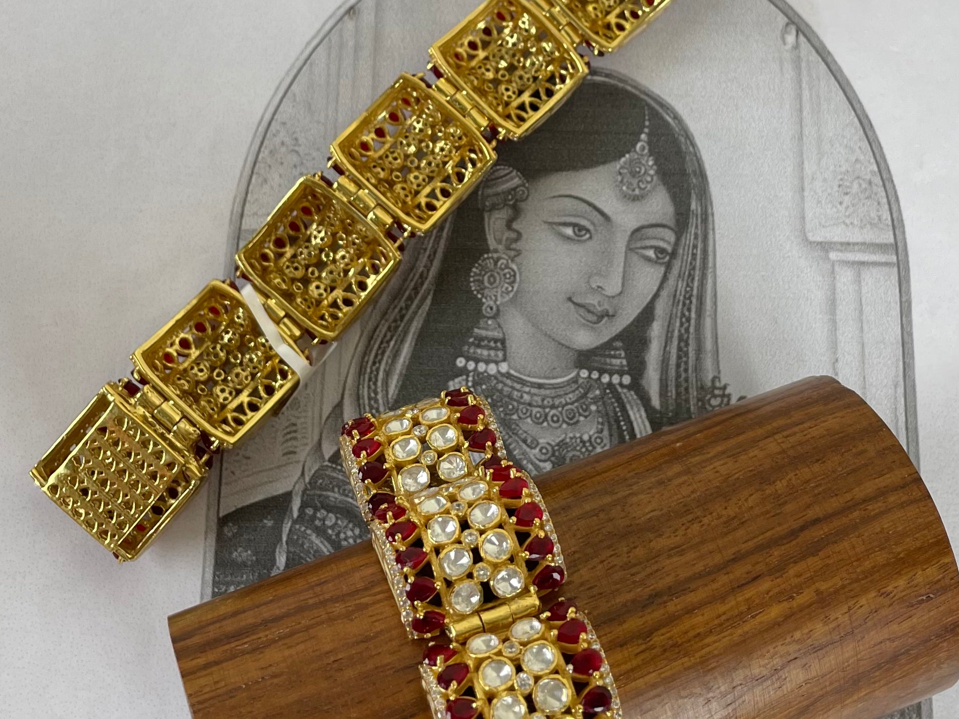 Gold-plated filigree bangles with red gemstone accents, displayed on a wooden stand in front of a vintage-style illustration of a woman.