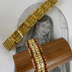 Gold-plated filigree bangles with red gemstone accents, displayed on a wooden stand in front of a vintage-style illustration of a woman.