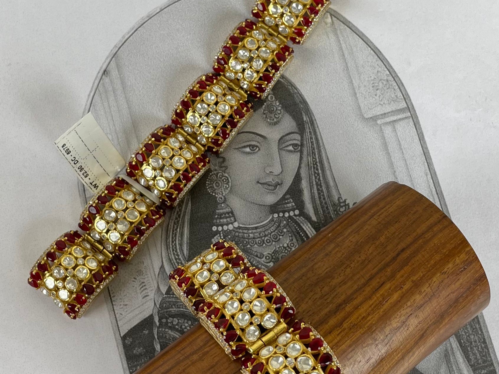 Two gold-plated Indian-style choker bracelets with red gemstone borders and clear central stones, displayed on a wooden stand over a vintage illustration card.
