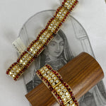 Two gold-plated Indian-style choker bracelets with red gemstone borders and clear central stones, displayed on a wooden stand over a vintage illustration card.