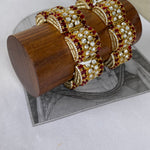 A set of ornate gold-toned bangles decorated with clear stones and deep red gemstones, displayed on a cylindrical wooden stand over a gray printed sheet.