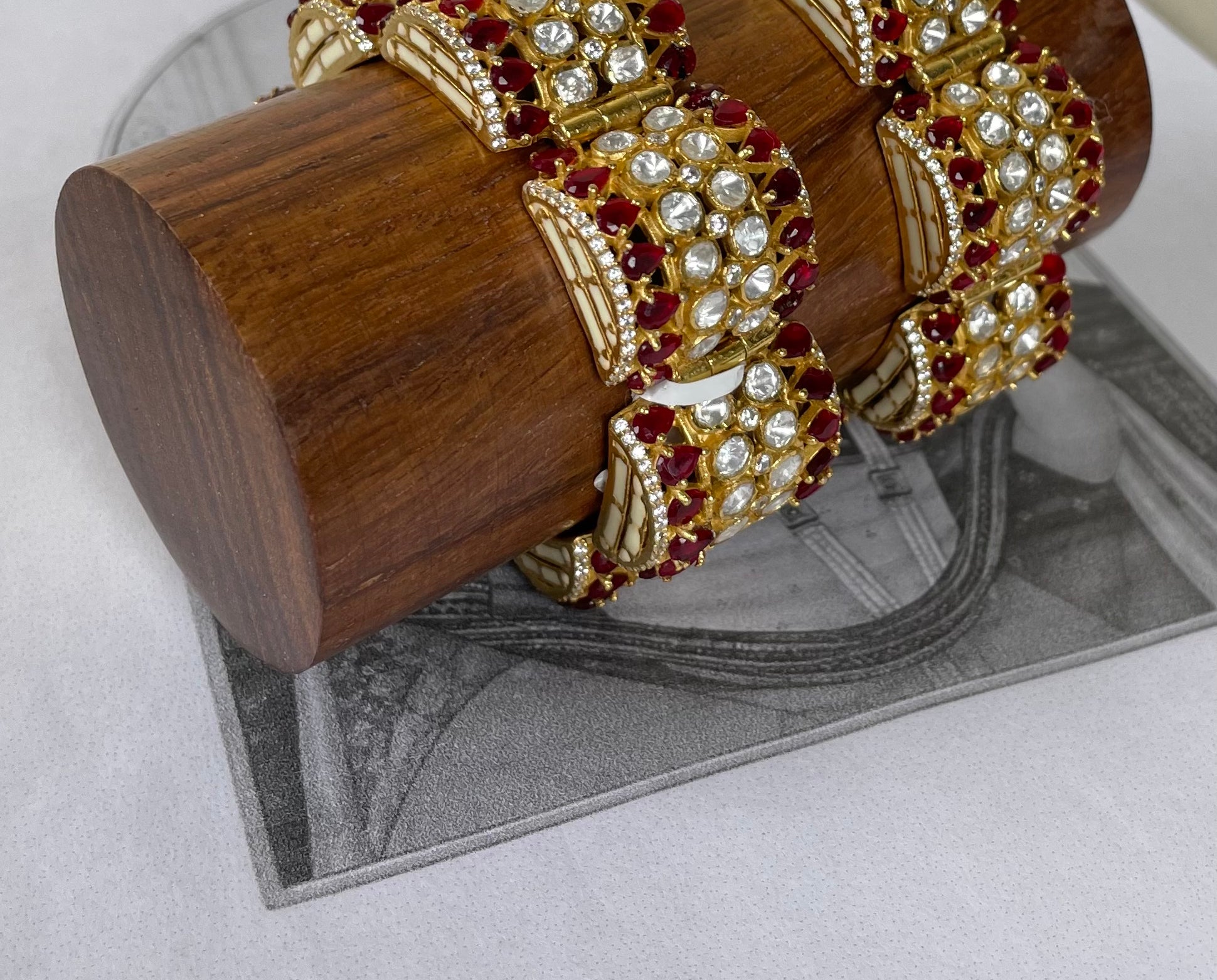 A set of ornate gold-toned bangles decorated with clear stones and deep red gemstones, displayed on a cylindrical wooden stand over a gray printed sheet.