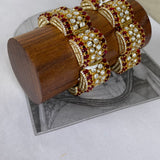 A set of ornate gold-toned bangles decorated with clear stones and deep red gemstones, displayed on a cylindrical wooden stand over a gray printed sheet.
