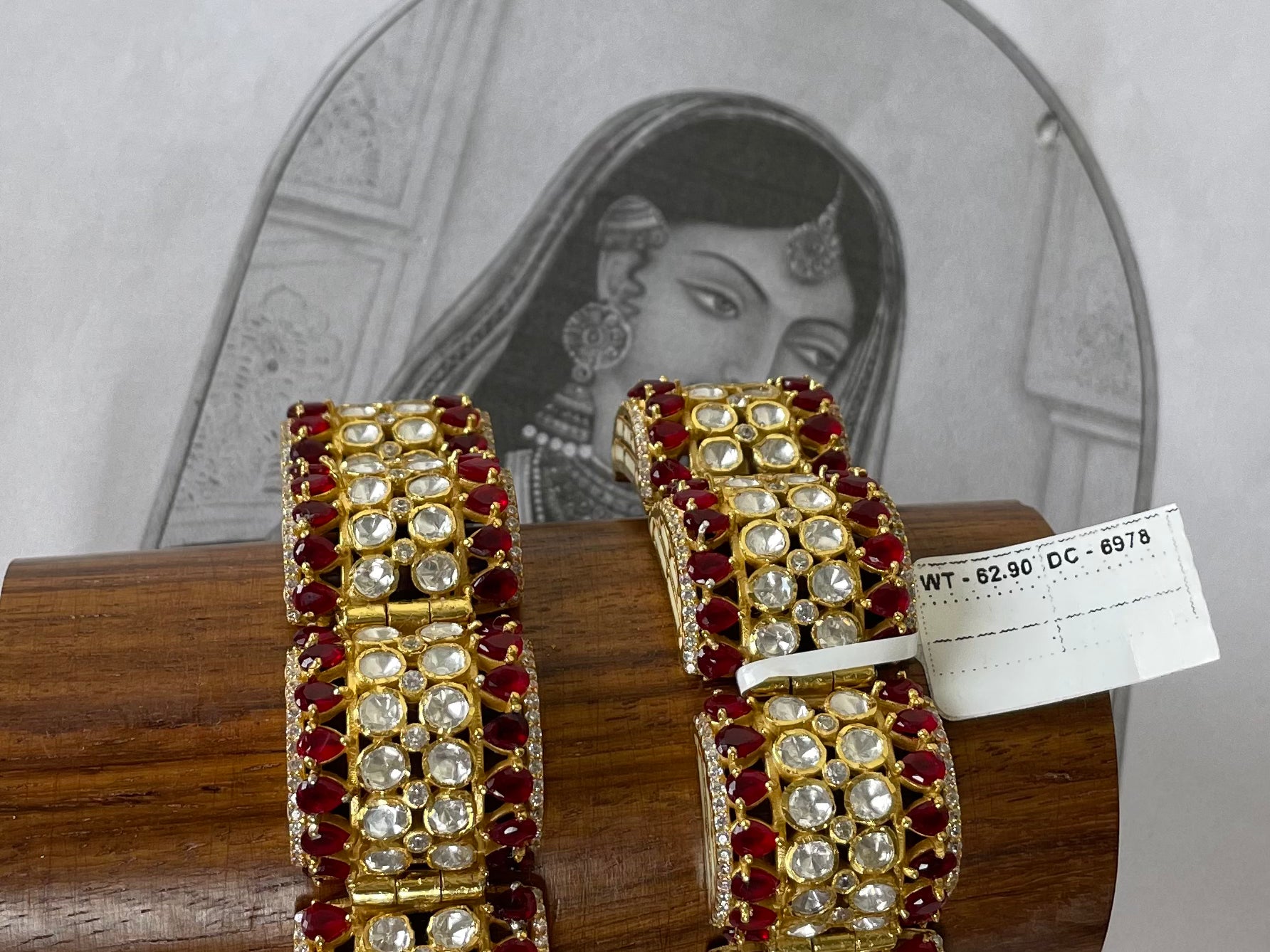 A pair of gold-plated Indian-style bangles decorated with red stones and white stones, displayed on a wooden stand in front of a vintage-style illustration.