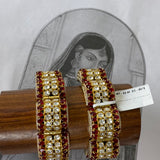 A pair of gold-plated Indian-style bangles decorated with red stones and white stones, displayed on a wooden stand in front of a vintage-style illustration.