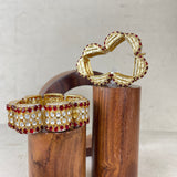 Two gold-toned statement bangles displayed on cylindrical wooden stands against a light textured background. One bracelet is a wide cuff set with clear stones and red gemstones, and the other is a flower-shaped bracelet with gold-toned petal segments outlined in red stones.