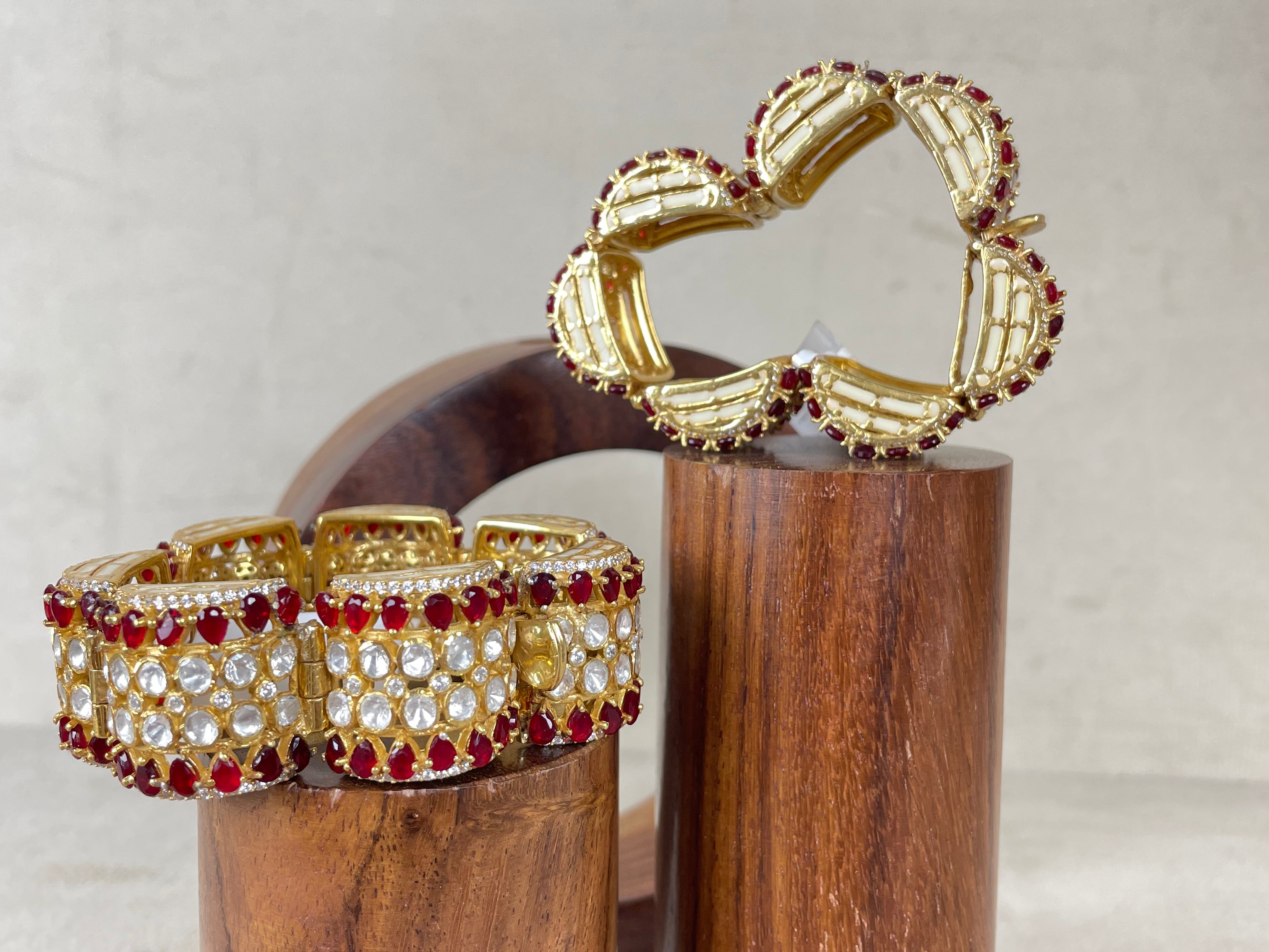 Two gold-toned statement bangles displayed on cylindrical wooden stands against a light textured background. One bracelet is a wide cuff set with clear stones and red gemstones, and the other is a flower-shaped bracelet with gold-toned petal segments outlined in red stones.