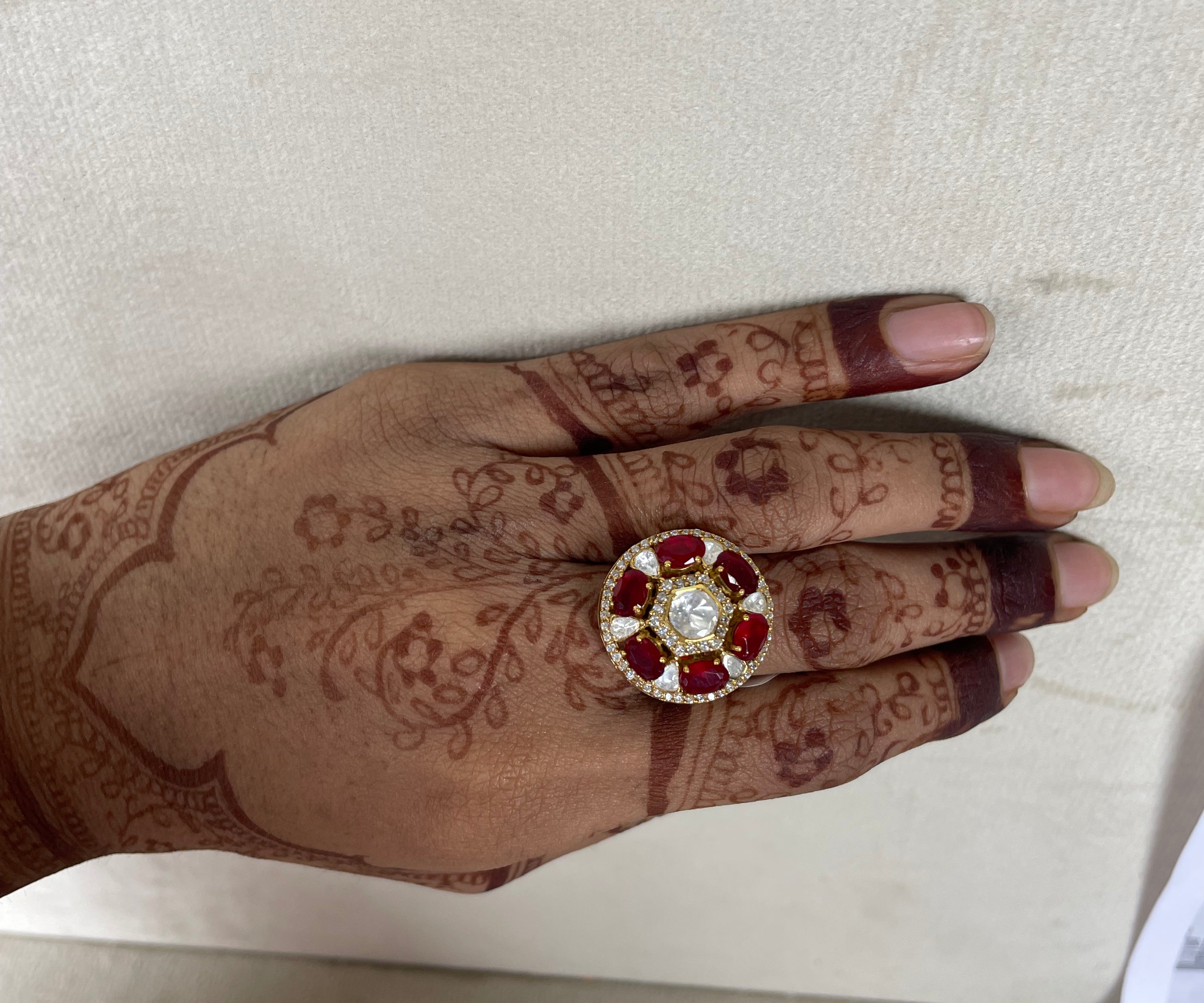 A close-up of a hand with intricate henna designs wearing a large circular ring. The ring features a central clear stone surrounded by red gemstones in a gold-toned setting, resting on a light beige textured surface.