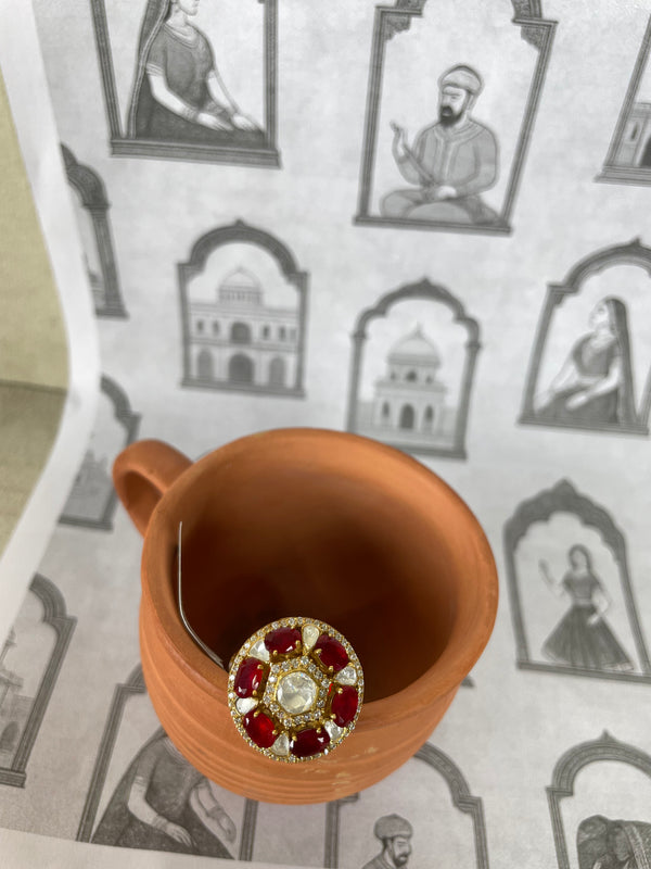 A terracotta-colored ceramic mug viewed from above, with a round gold-tone brooch featuring red gemstones and a central clear stone resting on the rim. The mug sits on patterned paper with black-and-white illustrations of figures and domed architecture.