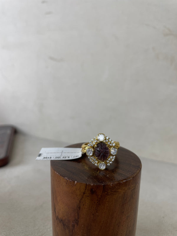 A vintage-style gold-plated ring with a central oval dark purple gemstone and surrounding clear stones, displayed on a wooden cylinder stand.