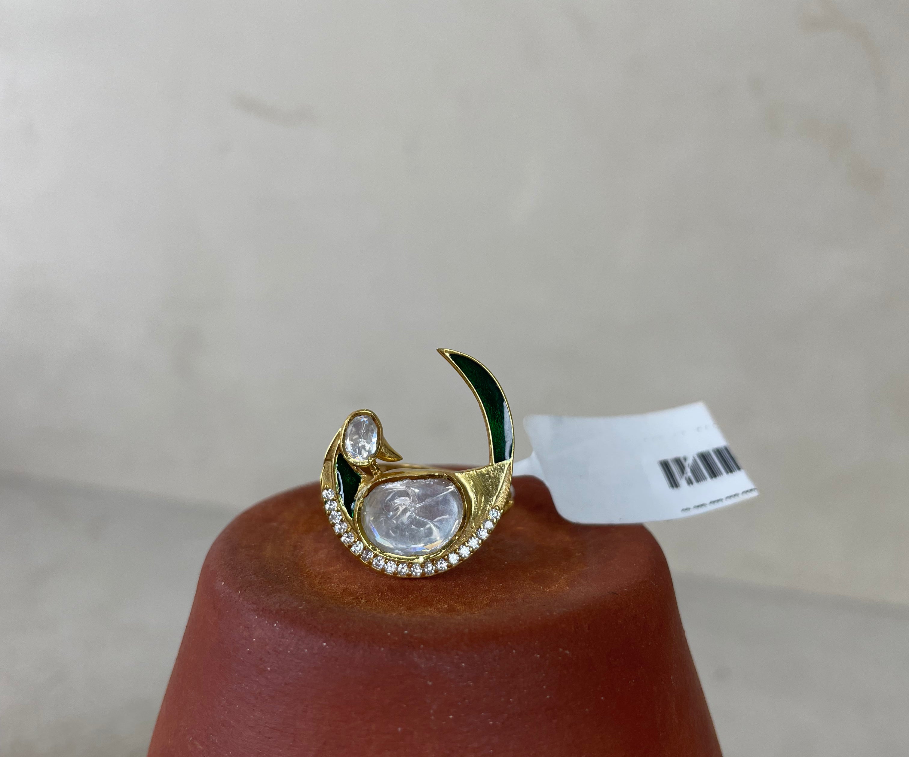 A gold crescent moon ring with a large clear teardrop gemstone, small clear stones, and dark green enamel accents, displayed on a terracotta cone with a white barcode tag attached.