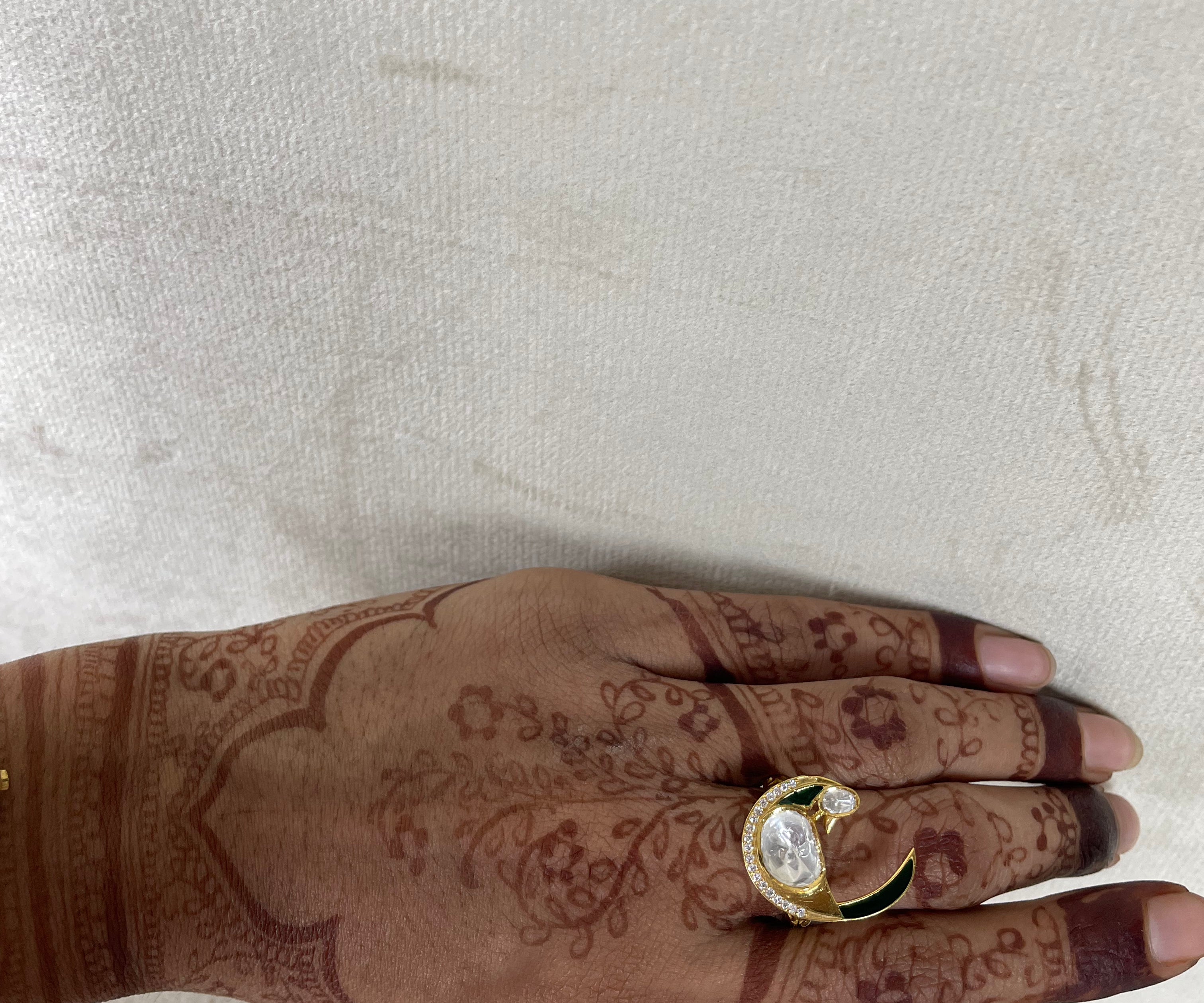 A gold-plated crescent moon ring with a clear central gemstone and green accents, worn on a hand decorated with brown henna designs, set against a textured off-white background.