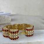 A gold-plated cuff bracelet adorned with rows of red gemstones and clear rhinestones, displayed on a light beige stone surface.