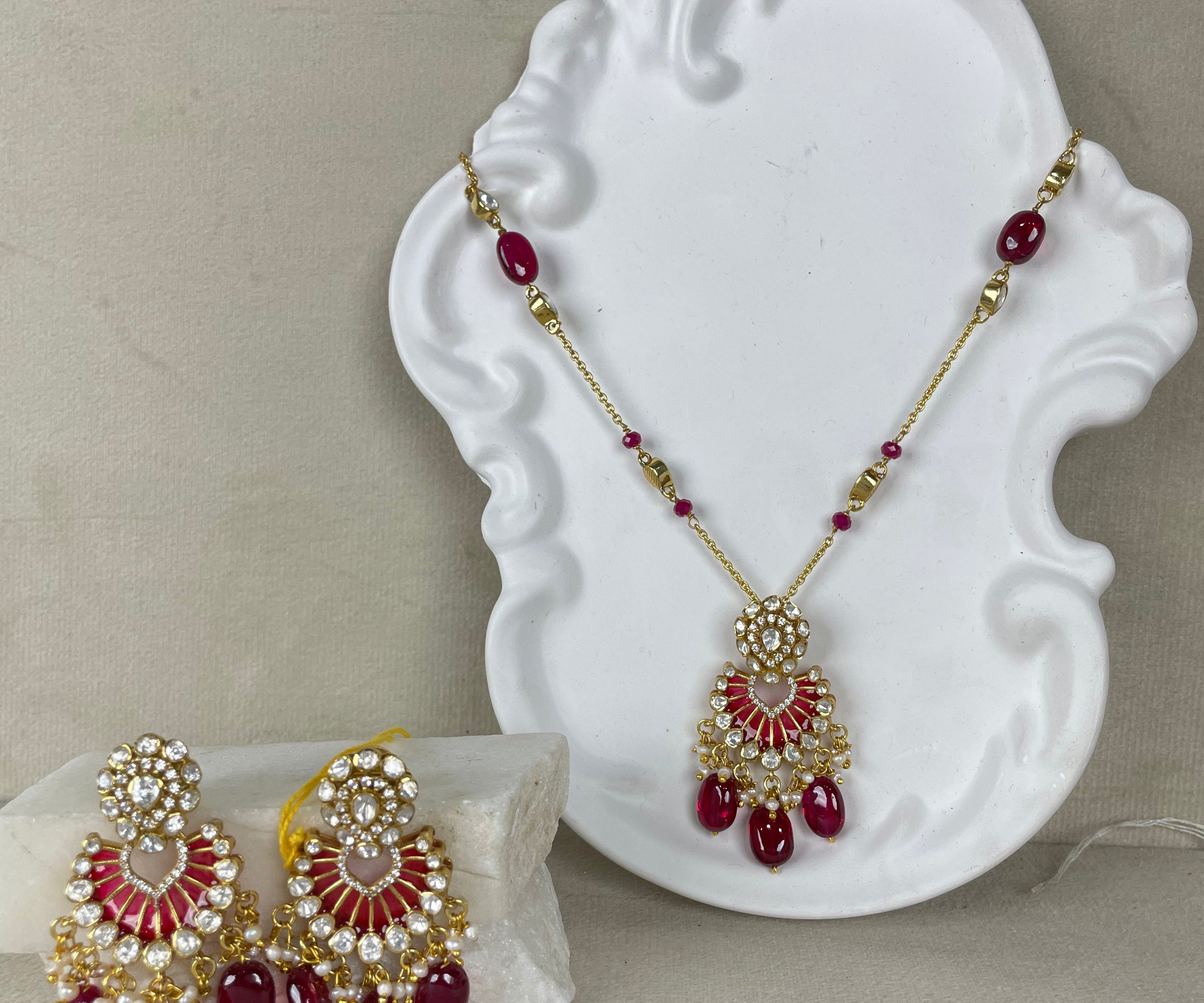 Gold-toned Indian jewelry set featuring a pendant and matching earrings with red enamel, clear stones, and red teardrop beads, displayed on a white decorative stand against a neutral beige background.