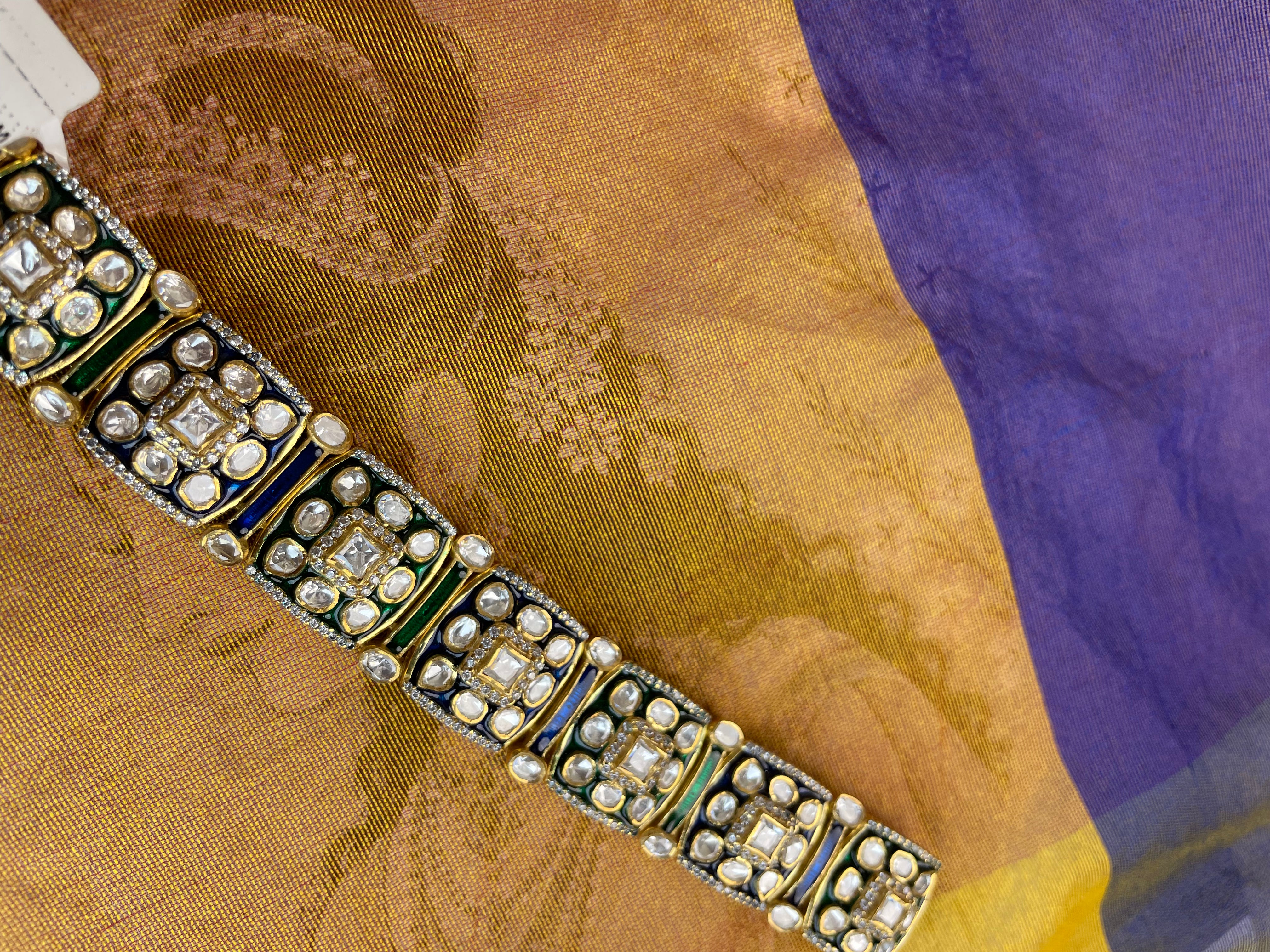 A close-up of an ornate embroidered sari fabric featuring a gold and purple body with a detailed metallic border embellished with silver zari work, blue enamel, and clear stones.