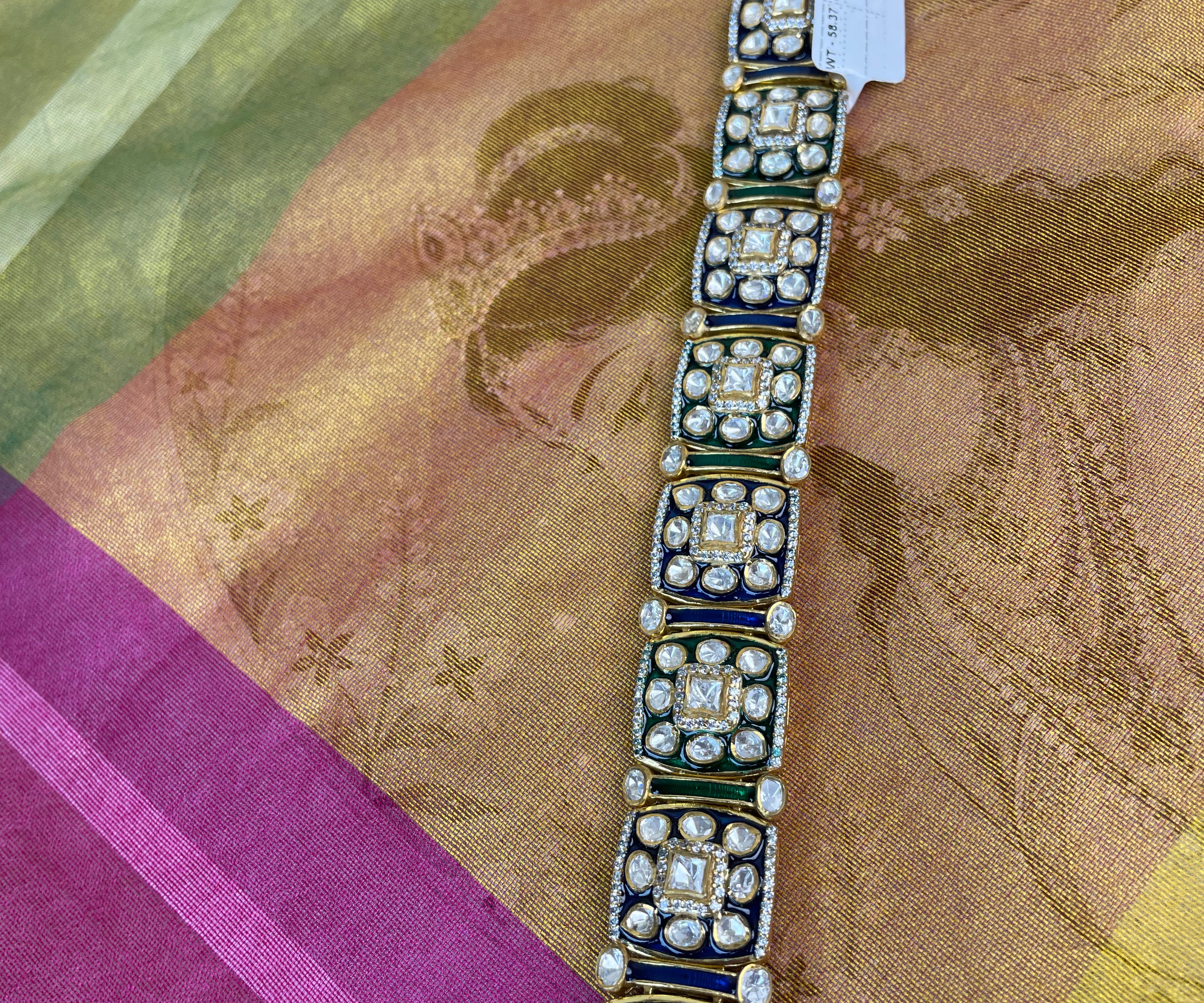 A gold-plated traditional Indian bracelet with rectangular links, decorated with green enamel, white stones, and a central red accent, laid flat on a patterned silk fabric.