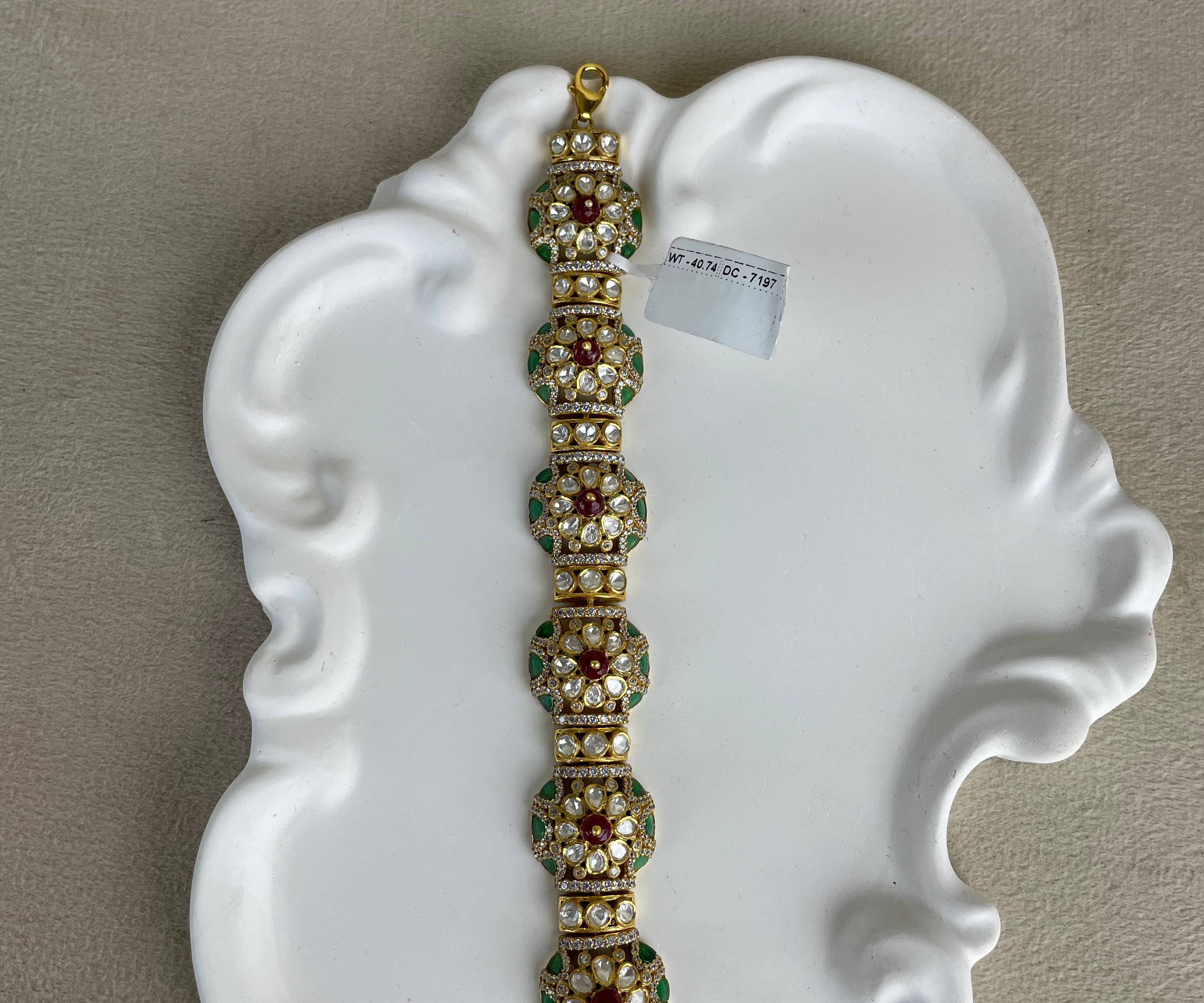 A gold-plated traditional Indian bracelet with green and red stone embellishments, displayed on a white decorative ceramic plate against a neutral beige background.