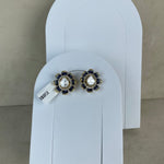 A pair of floral stud earrings with white pearl-like centers, dark blue stones, and gold-toned metal settings, displayed on white arch-shaped card holders against a beige wall.