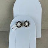 A pair of floral stud earrings with white pearl-like centers, dark blue stones, and gold-toned metal settings, displayed on white arch-shaped card holders against a beige wall.