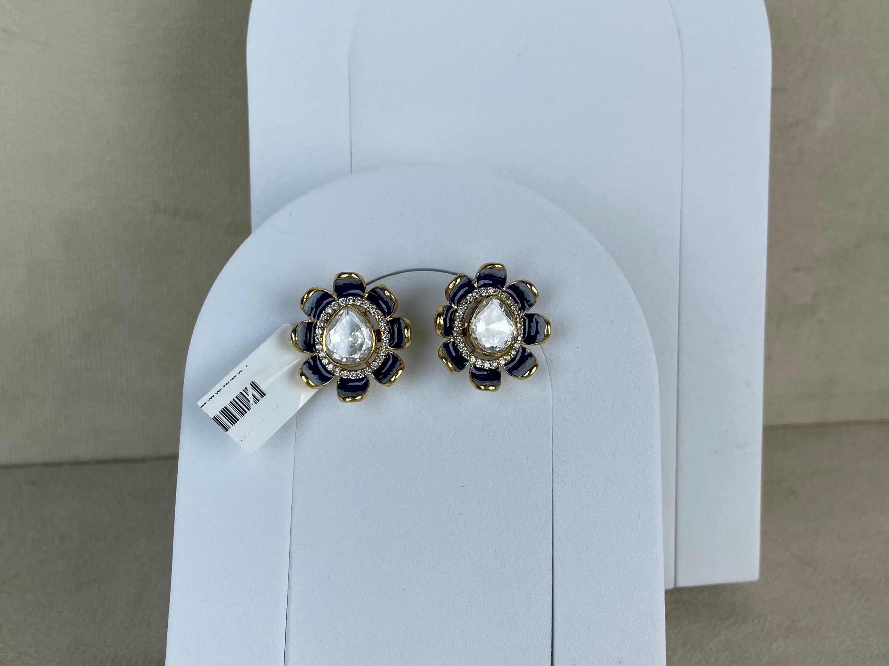 A pair of floral stud earrings with white pearl-like centers, dark blue stones, and gold-toned metal settings, displayed on white arch-shaped card holders against a beige wall.